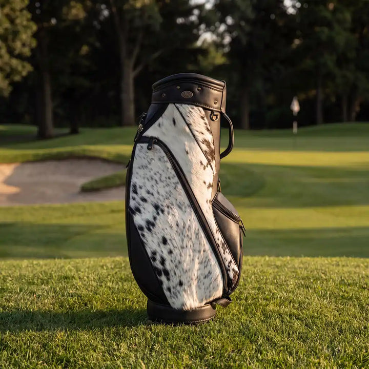 A golf bag with a black and white cowhide pattern and black leather trim, standing upright on a green grassy golf course.