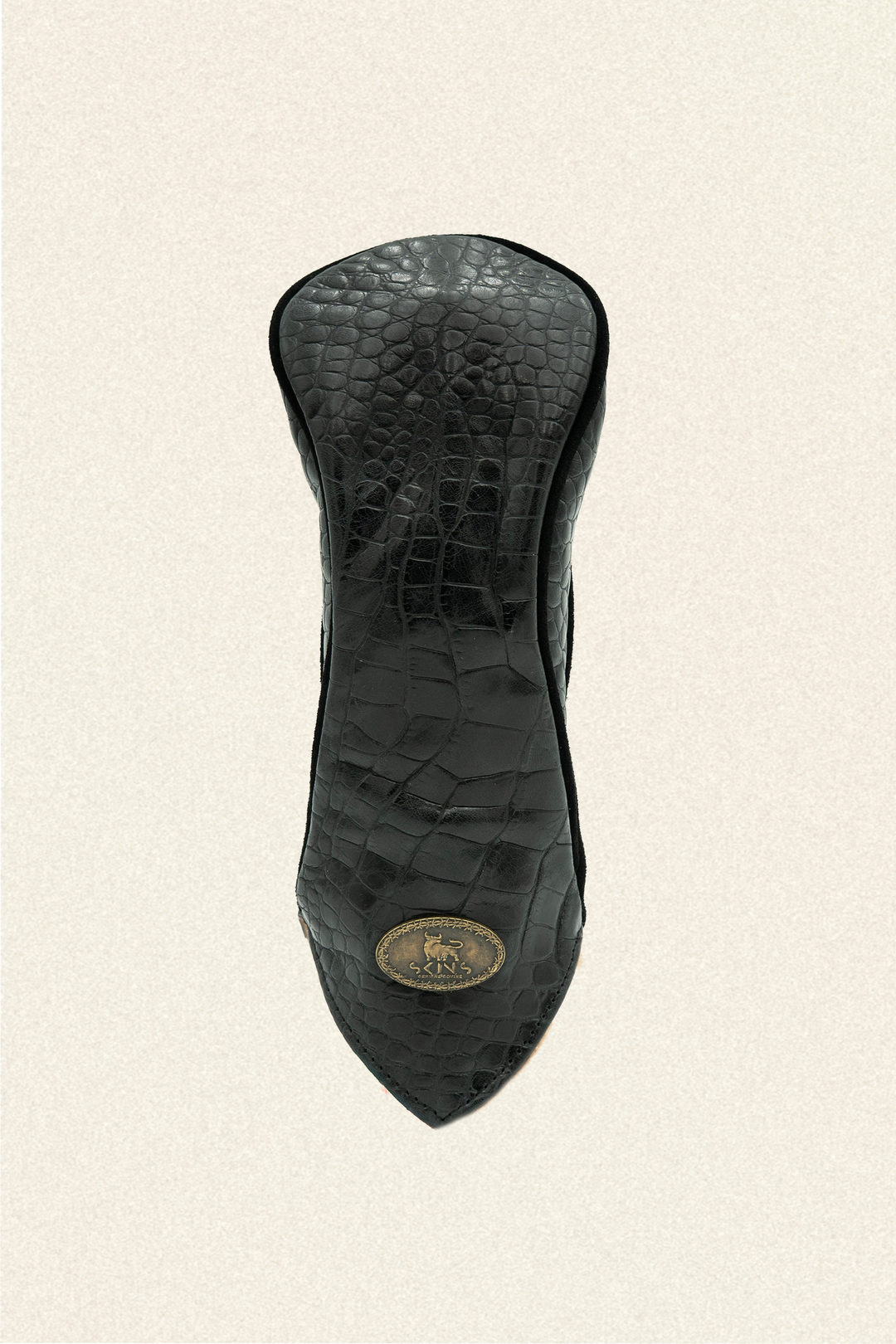 A black crocodile-embossed leather golf club headcover, shown from the back, featuring a gold-toned oval logo medallion near the pointed tip, set against a light beige textured background.