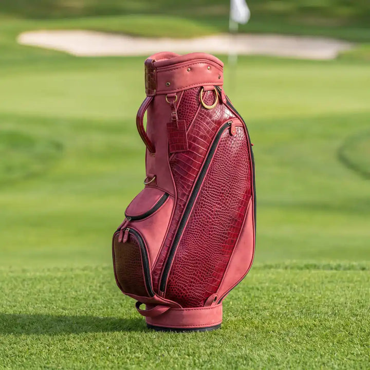 A red crocodile-embossed leather golf bag standing upright on a green golf course grass with a blurred background.