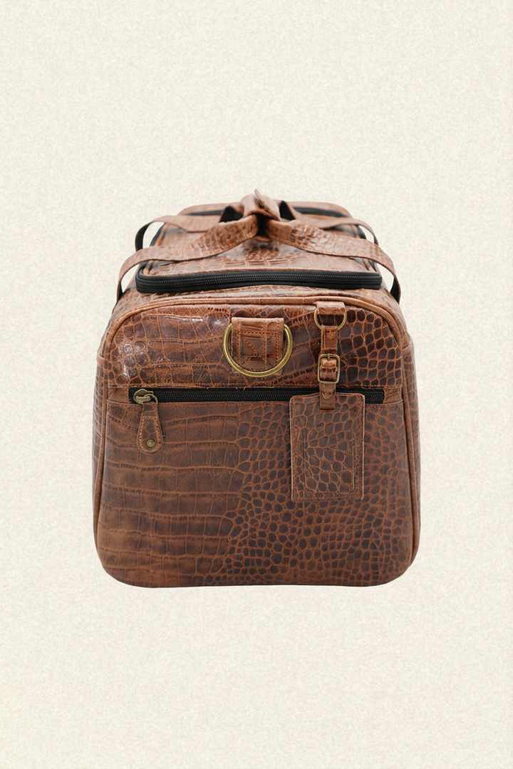 A brown crocodile-embossed leather duffel bag with brass hardware, shown from the front against a light beige textured background.