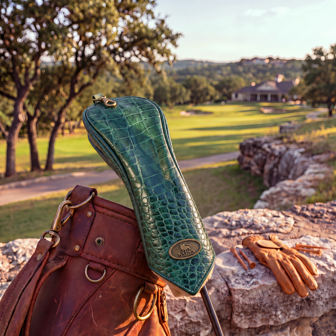 A green crocodile-embossed golf club headcover resting on a brown leather golf bag, with tan leather golf gloves on a stone wall and a blurred golf course background.