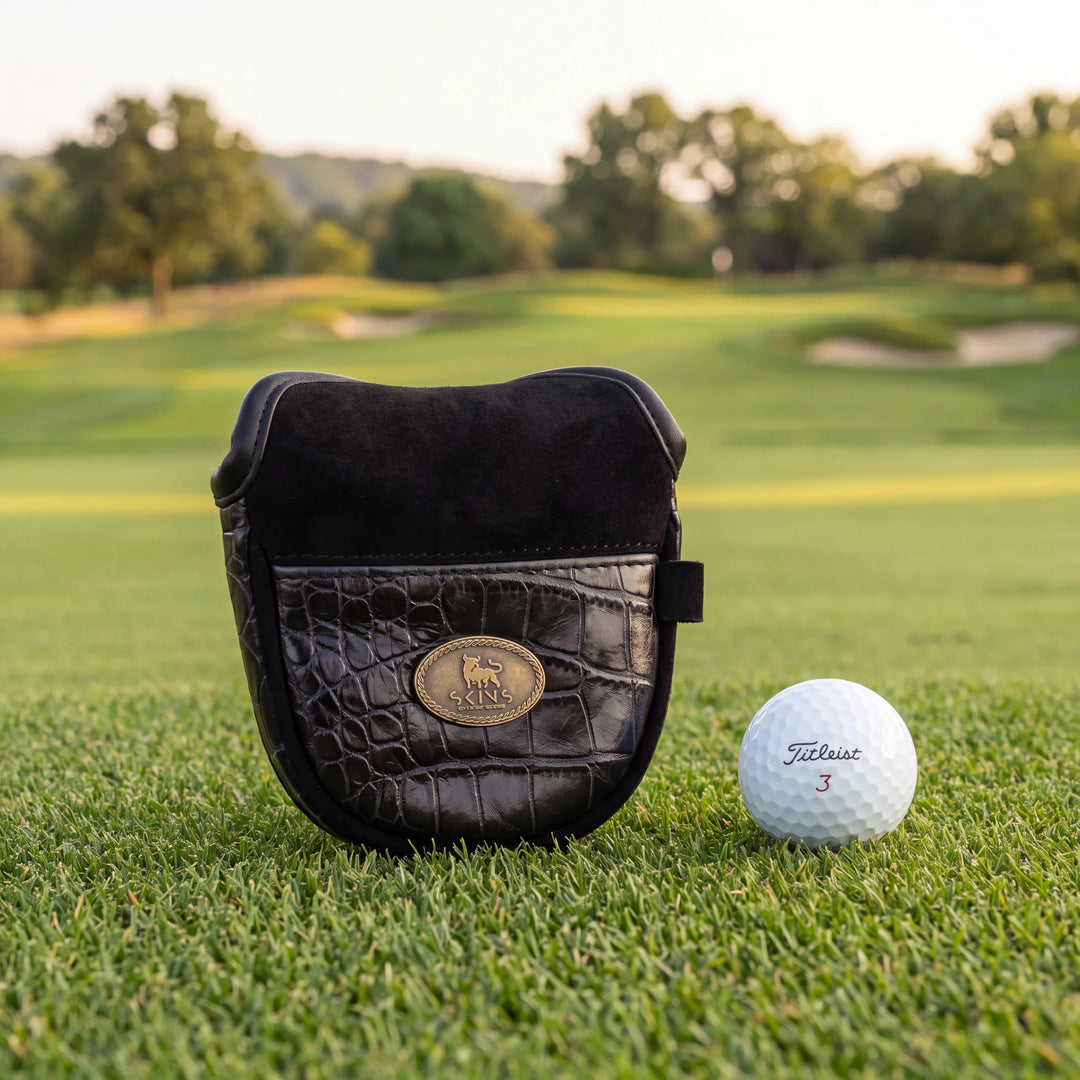 A black crocodile-embossed leather golf bag cover with a gold oval logo plaque sits on a green golf course next to a white Titleist golf ball.