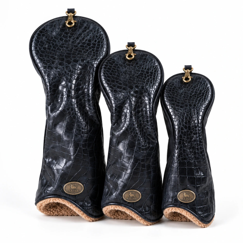 Three black crocodile-embossed leather golf club headcovers in graduated sizes, arranged side-by-side on a white background. Each cover features a gold-tone metal ring at the top and a circular gold logo badge near the bottom, with a tan textured lining visible at the base.