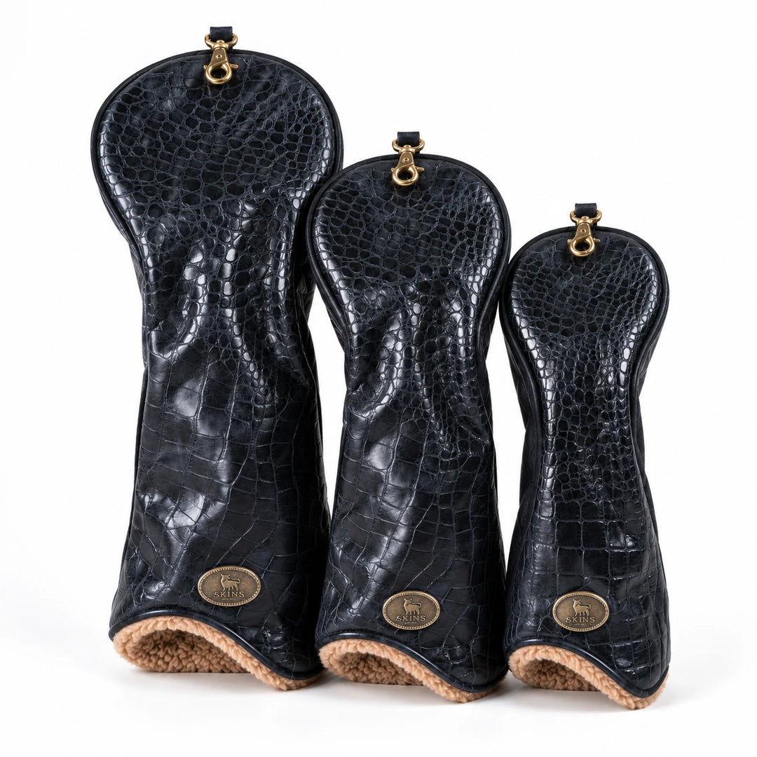 Three black crocodile-embossed leather golf club headcovers in graduated sizes, arranged side-by-side on a white background. Each cover features a gold-tone metal ring at the top and a circular gold logo badge near the bottom, with a tan textured lining visible at the base.