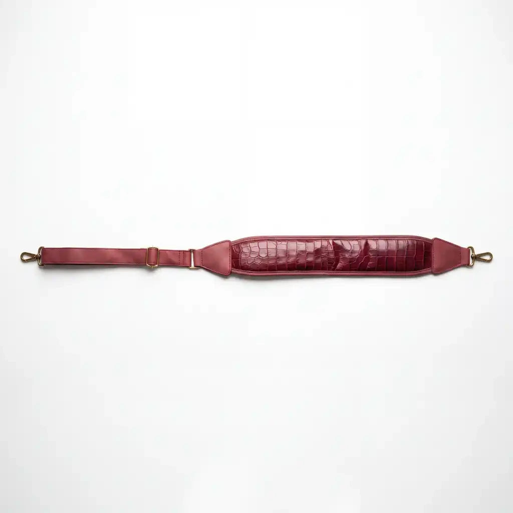 A burgundy leather shoulder strap with a crocodile-embossed texture on the padded section, brass-toned hardware, and an adjustable buckle strap, laid flat against a white background.