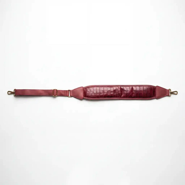 A burgundy leather shoulder strap with a crocodile-embossed texture on the padded section, brass-toned hardware, and an adjustable buckle strap, laid flat against a white background.