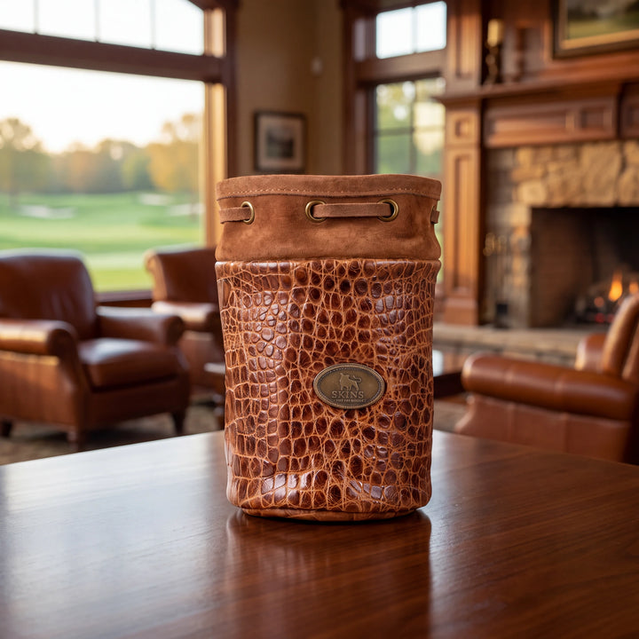 A brown crocodile-embossed leather bucket bag with a brass logo plaque, sitting on a polished wooden table in a warm, upscale living room with a fireplace and leather chairs in the background.