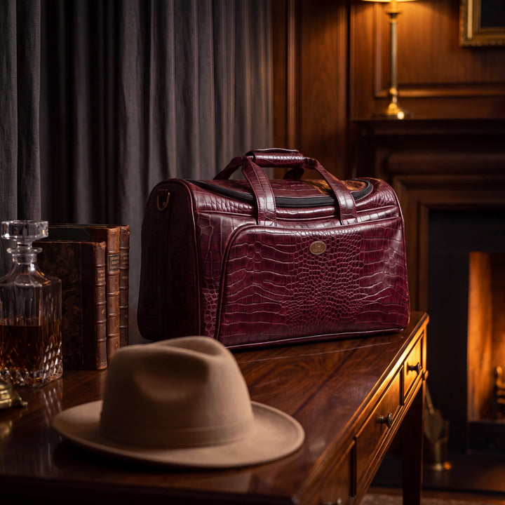 A burgundy crocodile-embossed leather duffel bag sits on a polished wooden table in a warm, darkly lit room. A beige fedora hat and a glass decanter are in the foreground, with books and a fireplace in the background.
