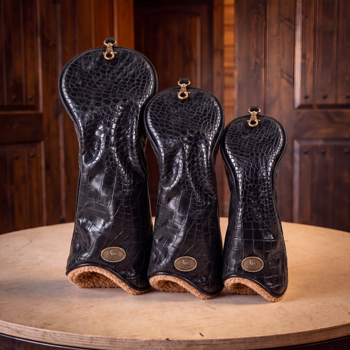 Three black crocodile-embossed leather golf club headcovers in different sizes, displayed on a round wooden table against a dark wood-paneled background.