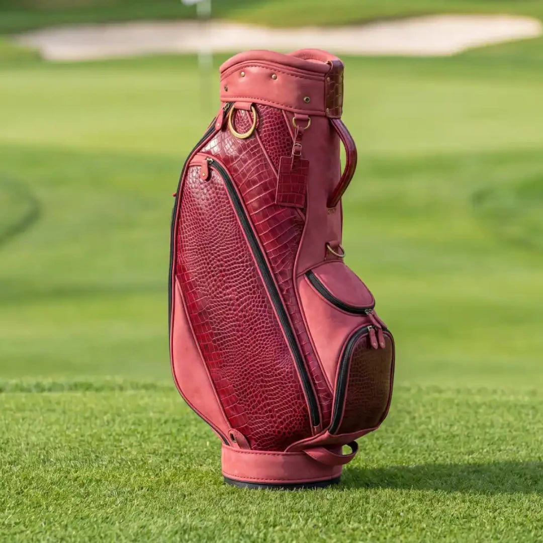 A pinkish-red leather golf bag with crocodile-embossed panels and black piping, standing on a green golf course.